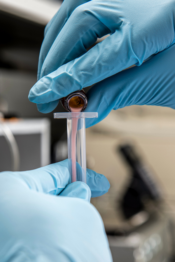 a close up image of hands wearing gloves pouring a pink substance into a test tube